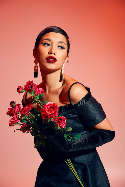 young and expressive asian woman with brunette hair, bold makeup, in black cocktail dress, long gloves and trendy earrings holding roses and looking away on pink background, stylish spring concept - Foto, afbeelding
