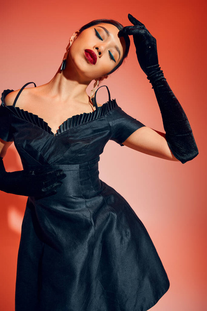 young and seductive asian woman with bold makeup, brunette hair, in black long gloves and cocktail dress posing with closed eyes on red and pink background, youth culture, trendy spring concept - Photo, Image