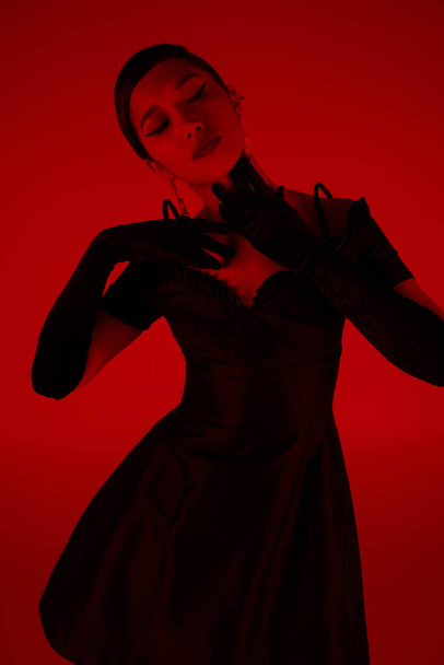 εκφραστική και χαριτωμένη Ασιάτισσα με μαύρα μακριά γάντια και κομψό φόρεμα κοκτέιλ αγγίζοντας το λαιμό και ποζάροντας σε ζωντανό φόντο με κόκκινο εφέ φωτισμού, ανοιξιάτικη φωτογραφία μόδας - Φωτογραφία, εικόνα