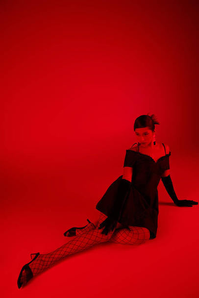 поколение z, весенняя концепция моды, полная длина молодой азиатской женщины в черном платье, длинные перчатки и трико рыболовных сетей сидя на ярком фоне с эффектом красного освещения - Фото, изображение