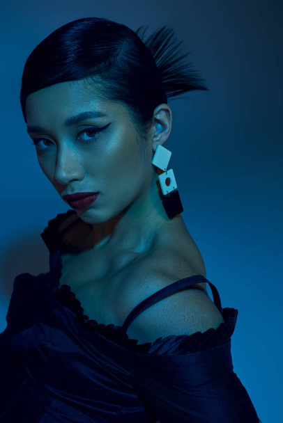 portrait of appealing asian woman with brunette hair, bold makeup, trendy earring and expressive gaze looking at camera on blue background with cyan lighting effect, youth culture, stylish spring  - 写真・画像