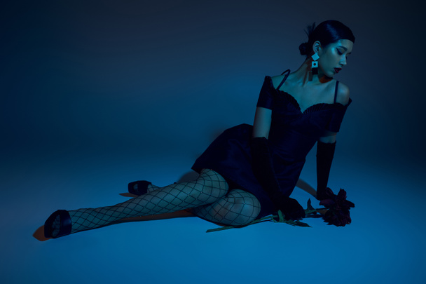 full length of sensual and elegant asian woman in black cocktail dress, long gloves and fishnet tights sitting near peony flower on blue background with cyan lighting effect, spring fashion concept - Foto, afbeelding