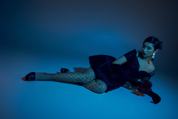 full length of graceful asian model in black cocktail dress, fishnet tights and long gloves laying near peony flower on blue background with cyan lighting effect, trendy spring, fashion shoot - Photo, Image