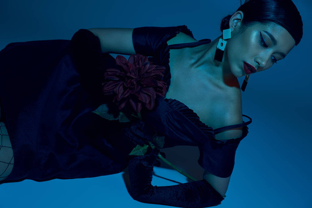 young and sensual asian woman in trendy earrings, black elegant dress and long gloves laying with peony flowers on blue background with cyan lighting effect, trendy spring, fashion shoot - Foto, Imagen