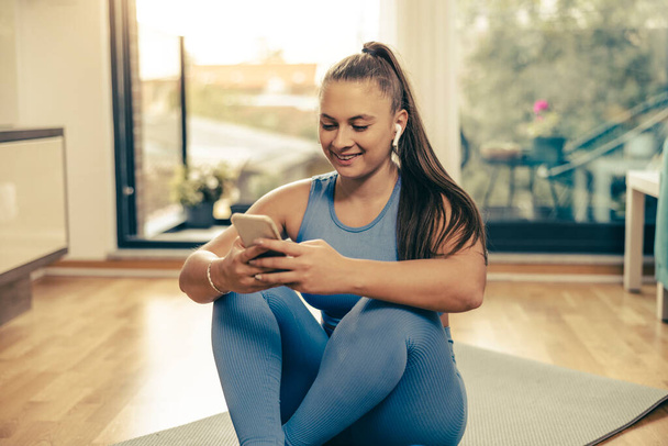 Giovane donna sorridente che ascolta musica dal suo smartphone e si prepara per l'allenamento a casa al mattino. - Foto, immagini