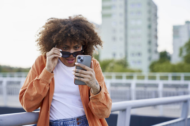 Черная женщина в солнечных очках и смотрит на свой мобильный телефон  - Фото, изображение