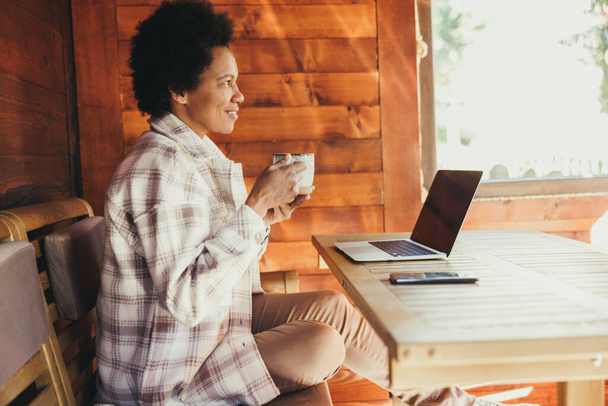 Mulher negra bebendo chá da manhã e usando laptop enquanto passa o tempo de férias em uma casa de madeira acolhedora. - Foto, Imagem