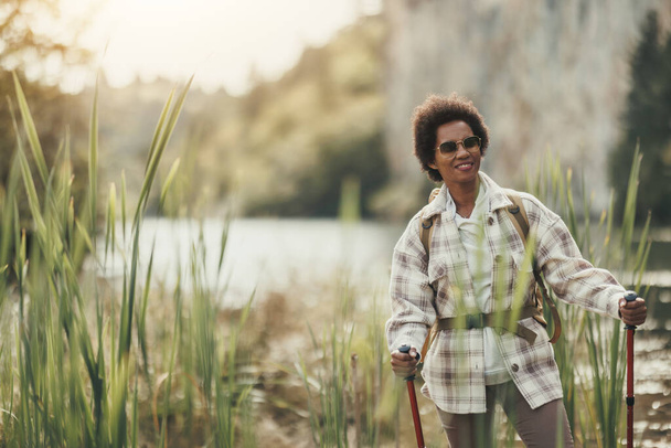 ゆったりとした大人の黒人女性で、川沿いにはリュックやトレッキングポールが立ち、山をハイキングしながらリラックスしています。. - 写真・画像