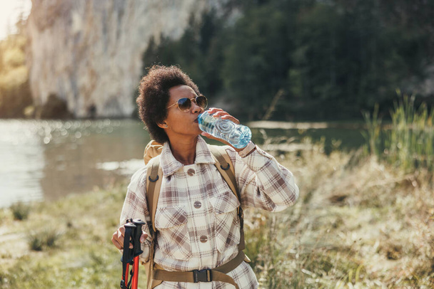 Ώριμη μαύρη γυναίκα με σακίδιο κάνοντας ένα διάλειμμα δίπλα στη λίμνη, ενώ έξω πεζοπορία στο βουνό. - Φωτογραφία, εικόνα