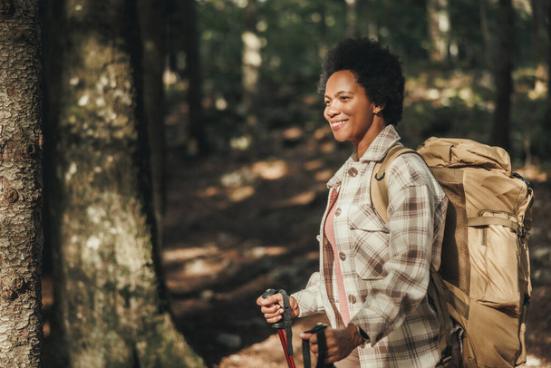 Smiling mature black woman standing alone outside and carrying a backpack while hiking in mountains. - Foto, imagen
