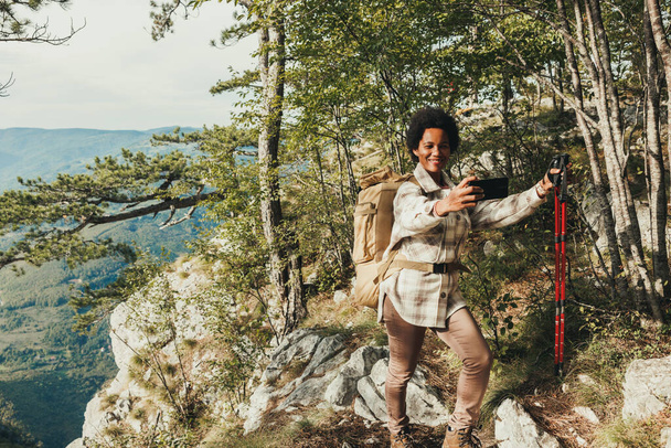 Mature black woman making a selfie while standing on the top a hill and enjoying the view during her hike in the mountains. - 写真・画像