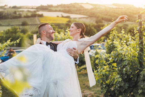 Cheerful groom carrying and spinning his bride while enjoying in vineyard on their wedding day. - Fotó, kép