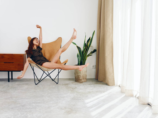 Mulher sentada em uma cadeira perto da janela sorrir alegres pés e mãos para cima, moderno estilo de vida escandinavo interior elegante, espaço de cópia. Foto de alta qualidade - Foto, Imagem