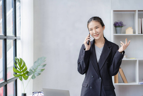 Привлекательная азиатская бизнесвумен разговаривает по телефону со своим деловым партнером в офисе с мобильным телефоном и ноутбуком - Фото, изображение
