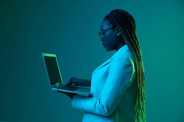 Afrikkalainen afrikkalainen nainen, jolla on kannettava tietokone laseissa sinisellä taustalla neonvalolla, värisekoitettu valo. Tietojen tallennus kyberturvallisuus, hakkeri, big data. Laadukas kuva - Valokuva, kuva