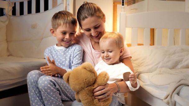 Gelukkig vrolijke jongens met moeder zitten op de vloer in de slaapkamer en spelen met teddybeer voor het naar bed gaan. - Foto, afbeelding