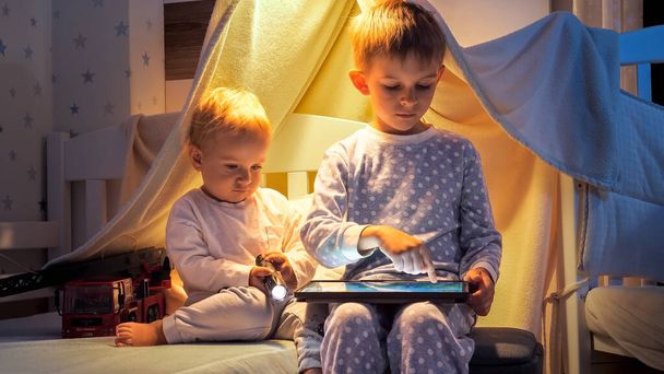 Dos chicos lindos en pijama jugando juegos en tableta en la cama por la noche. Niños con artilugios, educación, desarrollo infantil. - Foto, Imagen