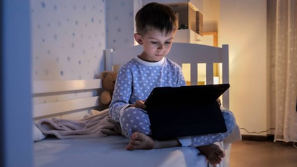 Pieni poika yöpuvussa, joka istuu tabletin kanssa sängyssä ja katsoo videota. Lapset koulutus, kehitys, lapset käyttävät vempaimia salaisuus, yksityisyys. - Valokuva, kuva