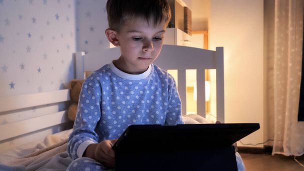 Portrait of boy in pajamas watching online video on tablet computer in bedroom at night. Children education, development, kids using gadgets secrecy, privacy. - Photo, Image