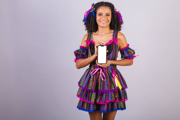 Black Brazilian woman wearing festa junina clothes. Fellowship of Saint John. Arraial, with smartphone, screen for ads and text. - 写真・画像