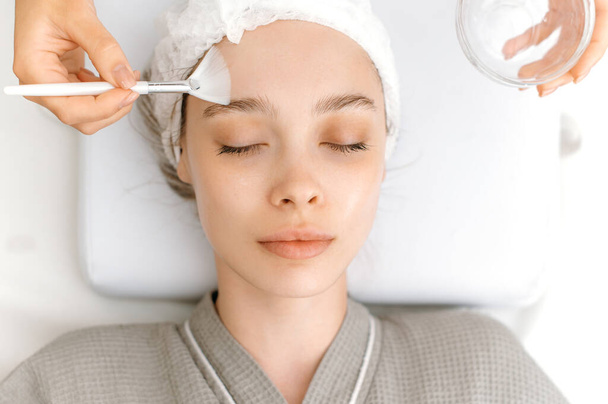 Kasvojen ihonhoitotoimenpiteet kauneushoitolassa. Kosmetologi puhdistaa kauniin valkoihoisen naisen kasvot erityisellä geelillä ennen maskin levittämistä, kuorimista, hankaamista. - Valokuva, kuva