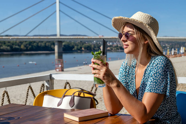 Una bella giovane donna che indossa un cappello di paglia e occhiali da sole, sorseggiando un cocktail e godendo di un bel pomeriggio estivo in un bar sulla spiaggia vicino al fiume. - Foto, immagini