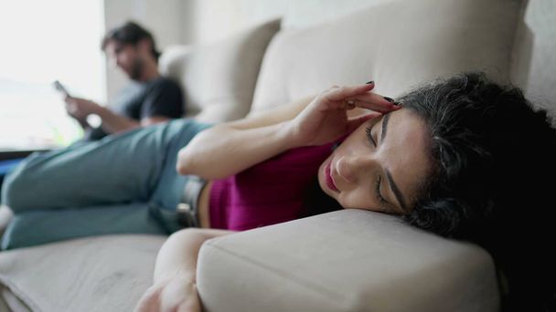 Jeune femme inquiète couchée sur un canapé souffrant de douleur émotionnelle. Femme en détresse dans les années 30 se sentant déconnecté de son petit ami en arrière-plan regardant son téléphone - Photo, image