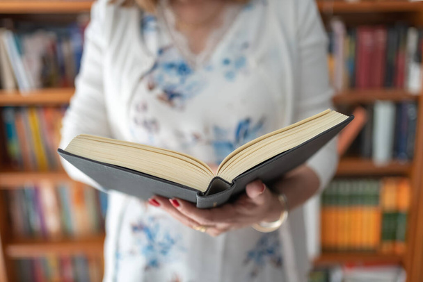Donna in piedi con un libro aperto mentre legge con una grande libreria dietro di lei - Foto, immagini