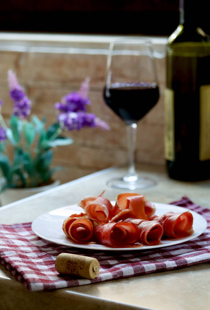 Plate with sliced prosciutto and a glass of red wine on kitchen table, lifestyle photography. Prsut and red wine, italian or spanish cured meats. Jamon appetizer with a glass of wine as brunch - 写真・画像