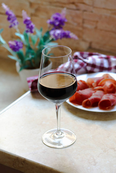 Glass of red wine and a plate with sliced prosciutto on kitchen table, lifestyle photography. Prsut and red wine, italian or spanish cured meats. Jamon appetizer with a glass of wine as brunch - Photo, Image