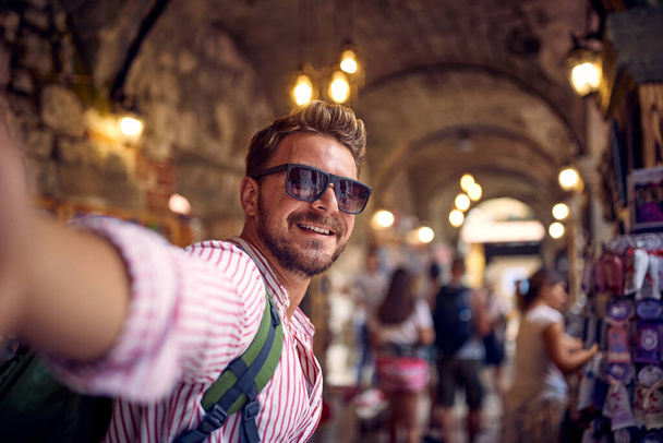Young man taking selfie in a street alley shop bazar. Tourist on vacation walking through street bazar. Holiday, fun, shopping, lifestyle concept. - Foto, imagen