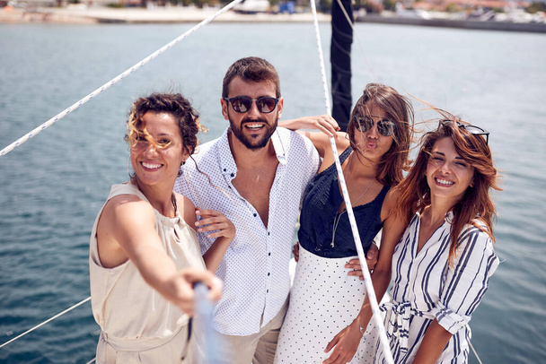A group of young handsome models is taking a selfie on the deck of the yacht while riding through the dock on a beautiful summer day on the seaside. Summer, sea, vacation, friendship - 写真・画像