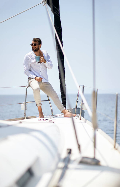 A young handsome male model at a photo shooting on a yacht on the seaside is enjoying a coffee and the sun on a beautiful sunny day. Summer, sea, vacation - Фото, зображення