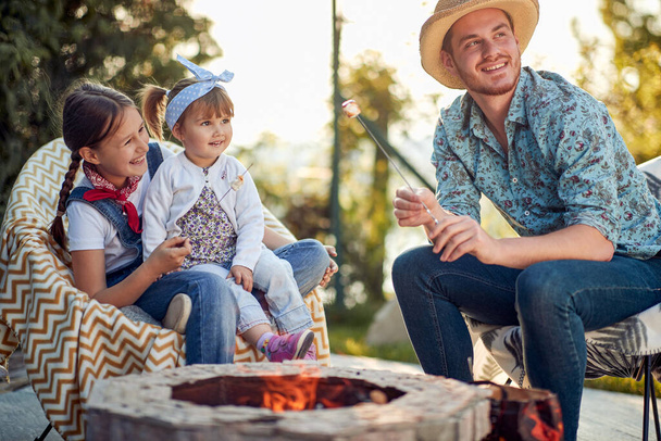 Joyful father roasting marshmallows with his two little daughters in a cozy cottage outdoors at daytime on a sunny summer day. Lifestyle, togetherness concept. - Foto, imagen