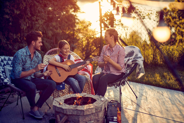 Little girl playing guitar by the fireplace, mom and dad clapping and singing along. Beautiful scene of family enjoying sunny summer day outdoors in the yard. Lifestyle, togetherness, family concept. - Photo, Image