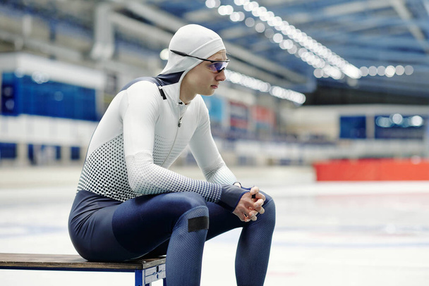 Seitenansicht eines jungen Sportlers, der auf einer Bank an der Eisbahn sitzt und sich nach hartem Training auf der Shorttrack-Eisschnelllaufbahn ausruht - Foto, Bild