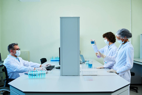 Side view of three coworkers in lab coats, protective masks and gloves working with new liquid substances obtained in laboratory - Photo, image