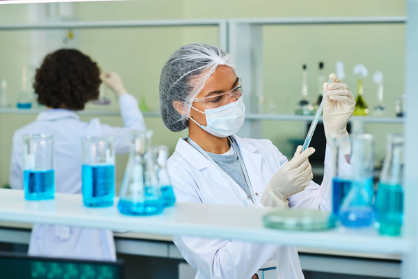 Junge Virologin oder Pharmakologin in der Hand eines Kolbens mit blauer flüssiger chemischer Substanz während eines wissenschaftlichen Experiments im Labor - Foto, Bild