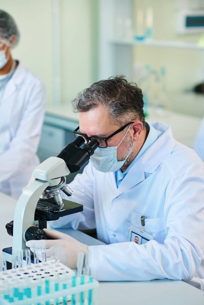 Volwassen mannelijke wetenschapper in laboratoriumjas, beschermende handschoenen en masker bestuderen van nieuwe stof in de microscoop terwijl zitten door de werkplek in het laboratorium - Foto, afbeelding