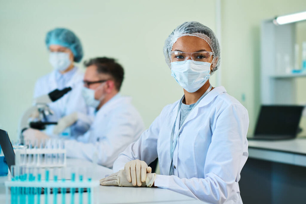 Mujer negra joven con bata de laboratorio, gorra médica, gafas protectoras, máscara y guantes mirando a la cámara mientras está sentada en el lugar de trabajo en el laboratorio - Foto, Imagen