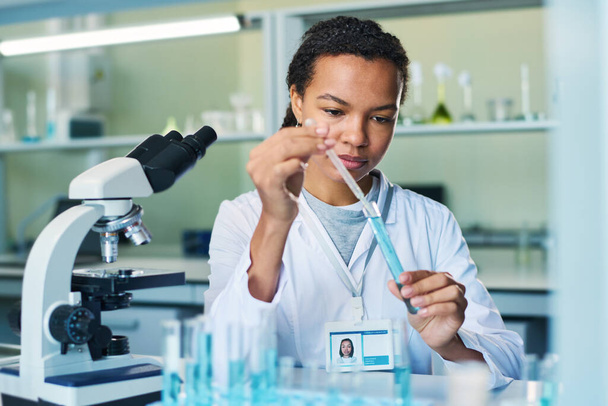 Νεαρή γυναίκα φαρμακοποιός με σταγονόμετρο που λαμβάνει δείγμα μπλε υγρής ουσίας από τη φιάλη κατά τη διάρκεια κλινικού πειράματος σε εργαστήριο - Φωτογραφία, εικόνα