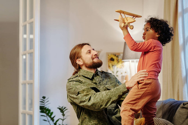 Joyeux jeune homme en uniforme militaire s'amusant avec son mignon petit fils jouant avec un avion jouet tout en se tenant devant lui à la maison - Photo, image