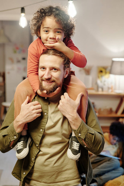 Happy young man looking at camera while standing in living room and holding his adorable little son on shoulders while having fun - Photo, Image