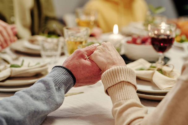 Χέρια στοργικού ηλικιωμένου ζευγαριού προσεύχονται σερβίροντας γιορτινό τραπέζι με σπιτικό φαγητό και αναμμένα κεριά πριν το δείπνο των Ευχαριστιών - Φωτογραφία, εικόνα