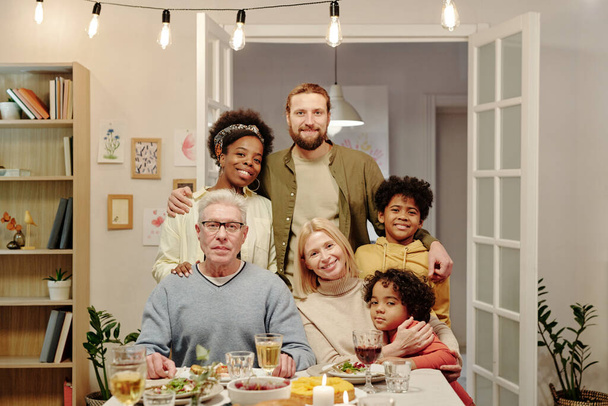 Duża wesoła międzykulturowa rodzina trzech pokoleń patrząc w kamerę siedząc naprzeciwko serwowanego stołu i ciesząc się świąteczną kolacją - Zdjęcie, obraz