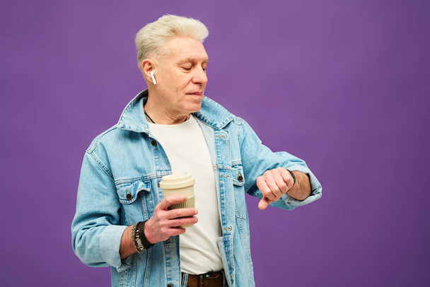 Starší sebevědomý muž v džínové bundě a bílém tričku při pohledu na hodinky a při čekání na někoho s kávou - Fotografie, Obrázek