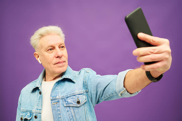 Selbstbewusster älterer Mann mit grauen Haaren hält Smartphone in der Hand, während er während der Online-Kommunikation den Arm ausstreckt oder ein Selfie macht - Foto, Bild