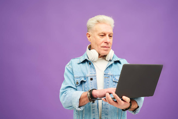 Zelfverzekerde senior zakenman met grijs haar typen op laptop toetsenbord tijdens het netwerken tegen lila achtergrond in isolatie - Foto, afbeelding