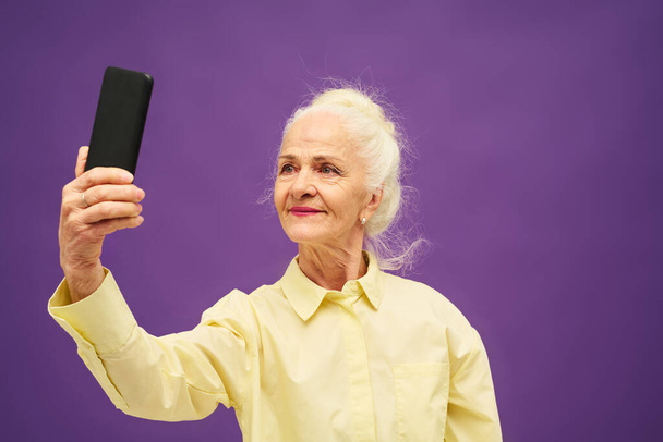 Senior smiling woman in yellow shirt looking at smartphone screen while taking selfie or communicating in video chat over violet background - Фото, изображение