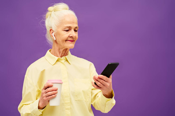 Senior vrouw met kopje koffie kijken naar online video in mobiele telefoon terwijl staan voor de camera over violette achtergrond - Foto, afbeelding